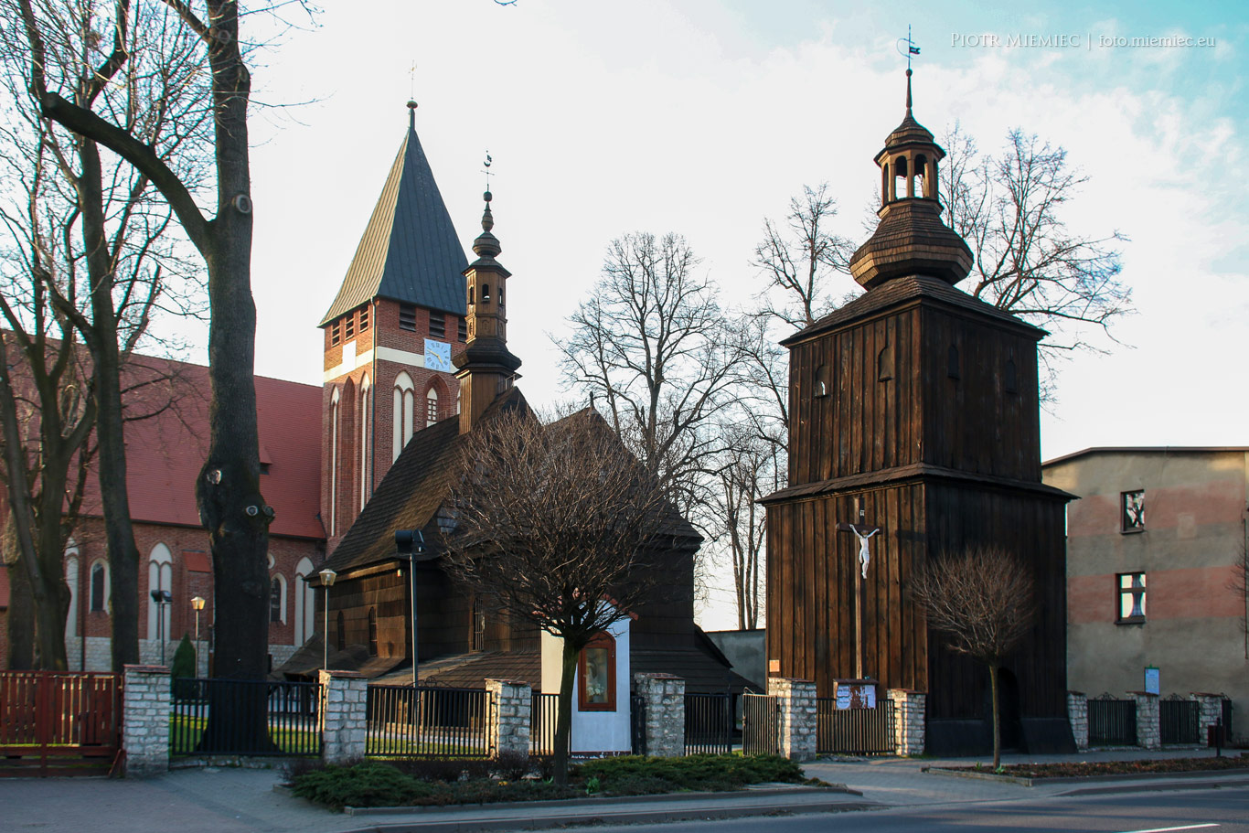 Zestawienie trzech świątyń w Miasteczku Śląskim: na pierwszym planie masywna drewniana dzwonnica z 1717 r., obok drewniany kościółek, a w tle wysoka murowana świątynia z czerwonej cegły.