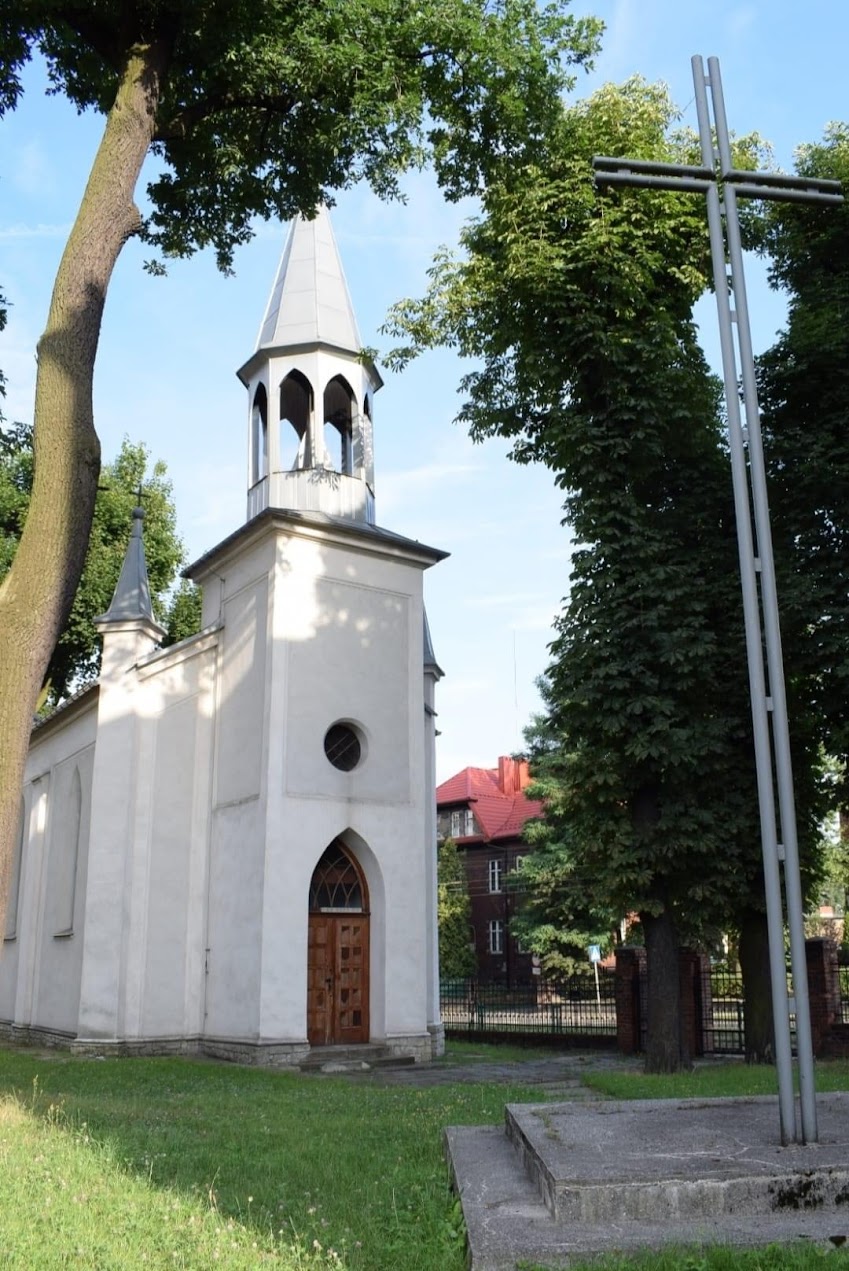 Współczesne zdjęcie białej, zabytkowej kaplicy w Bytomiu-Szombierkach, ujęcie od frontu z widoczną wieżą, drewnianymi drzwiami i wysokim krzyżem.