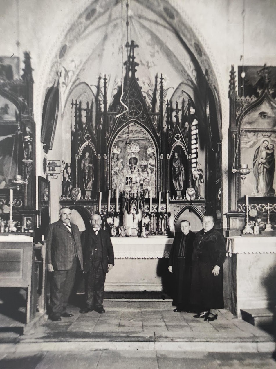 Historyczne zdjęcie wnętrza neogotyckiej kaplicy w Szombierkach. Przed bogato zdobionym ołtarzem stoją dwaj mężczyźni i dwie kobiety w eleganckich strojach z epoki.