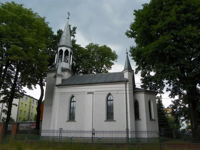 Widok z boku na murowaną, otynkowaną na biało kaplicę z ostrołukowymi oknami, otoczoną drzewami i metalowym płotem.